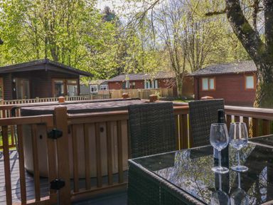 A deck with a hot tub and two chairs around a glass table with wine glasses and a bottle at Robins Lodge White Cross Bay near Troutbeck Bridge