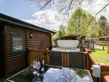 An outdoor patio with a glass table two wine glasses and a bottle next to a wooden cabin and a hot tub at Robins Lodge in White Cross Bay near Troutbeck Bridge