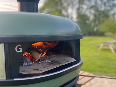 An outdoor wood fire oven with flames inside and a green lawn with a picnic table in the background at Pippin lodge in Coxbridge near Glastonbury