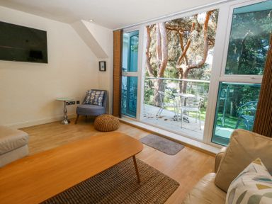 A living room with a sofa, wooden coffee table, armchair, side table, wall-mounted TV, and glass doors opening to a balcony with metal chairs and table at 6 Red Sails in Sandbanks