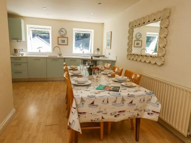 A kitchen with green cabinets two windows a dining table with a boat patterned tablecloth and six wooden chairs at 6 Red Sails in Sandbanks