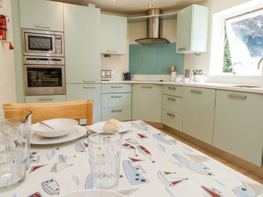 A kitchen with light green cabinets a built-in oven and microwave a wooden chair and a table with a tablecloth with boat prints at 6 Red Sails in Sandbanks