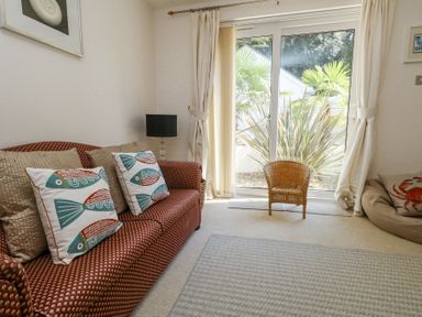 A living room with a patterned sofa and fish design cushions near sliding glass doors with a wicker chair and plants outside at 6 Red Sails in Sandbanks