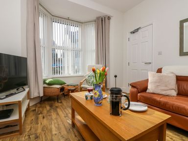 A living room with a brown leather sofa a wooden coffee table with a French press and flowers a white armchair a television and a bay window with vertical blinds at Breakers Point in St Ives
