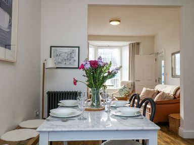 A dining table set with plates glasses and flowers in a vase with a living room and sofa in the background at Breakers Point in St Ives