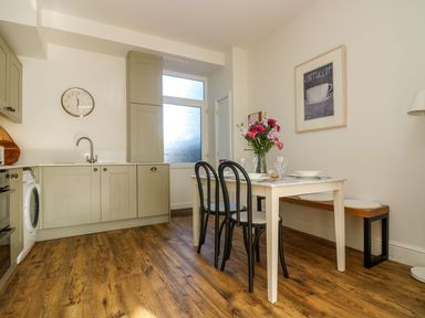 A kitchen with green cabinets a clock on the wall a dining table with flowers plates and glasses and black chairs at Breakers Point in St Ives