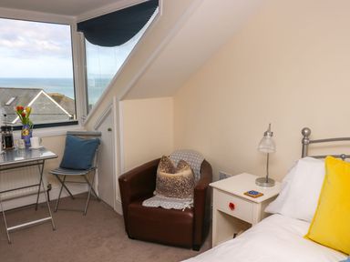 A bedroom corner with a bed yellow pillow bedside table lamp brown armchair with pillows small table and chair near window showing sea view at Breakers Point in St Ives