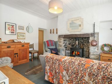 A living room with a wood stove fireplace an armchair a wooden cupboard a patterned sofa and framed pictures on the wall at Cobweb Cottage in Waunfawr