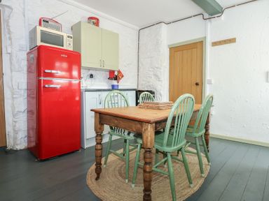 A kitchen with a wooden dining table surrounded by four green chairs a red refrigerator and a microwave on white walls at Cobweb Cottage in Waunfawr