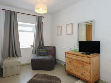 A small room with a window covered by gray curtains a plaid-covered chair a wooden chest of drawers with a TV on it and a mirror on the wall at Cobweb Cottage in Waunfawr