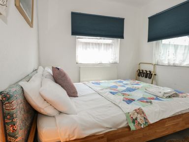 A bedroom with a bed covered with white sheets and a multicolored quilt under two windows with blue blinds at Cobweb Cottage in Waunfawr