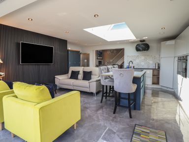 An open living room and kitchen area with yellow chairs a sofa bar stools and a wall-mounted television at Bronallt in Cemaes Bay