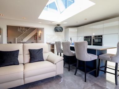 A kitchen with bar stools around an island and a beige sofa with cushions in a living area at Bronallt in Cemaes Bay