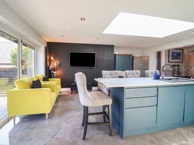 A kitchen and living area with yellow armchairs a blue island with gray chairs and a black textured wall with a mounted TV at Bronallt in Cemaes Bay