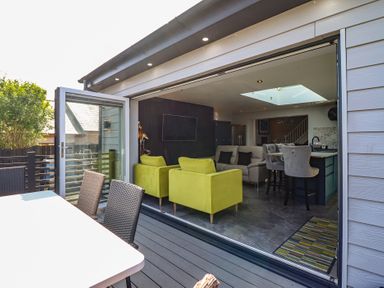 An open living area with green armchairs a sofa and bar stools visible from an outdoor deck with a table and chairs at Bronallt in Cemaes Bay