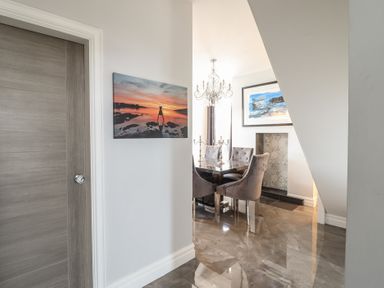 A hallway leading to a dining area with a table, five upholstered chairs, a chandelier, two paintings on the walls, and a fireplace at Bronallt in Cemaes Bay