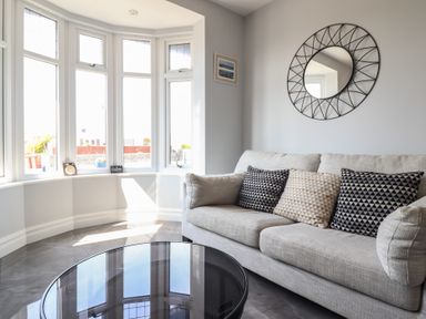A living room with a beige sofa with patterned cushions and a round glass coffee table near a bay window at Bronallt in Cemaes Bay