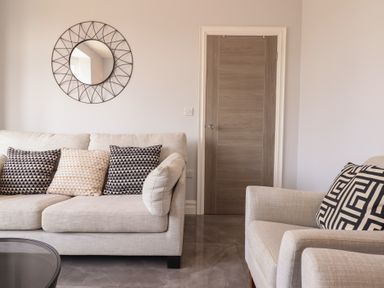 A living room with a beige sofa and armchair with patterned cushions a round wall mirror and a wooden door at Bronallt in Cemaes Bay