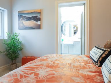 A bedroom with an orange bedspread and pillows beside a plant and a painting with a door leading to a bathroom at Bronallt in Cemaes Bay