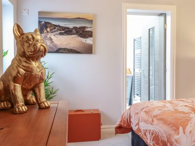 A bedroom with a bed covered by a patterned duvet a wooden dresser with a gold dog statue a landscape picture on the wall and an open door leading to a bathroom at Bronallt in Cemaes Bay