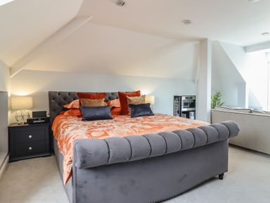 A bedroom with a grey upholstered bed orange bedding two bedside tables with lamps and a sitting area at Bronallt in Cemaes Bay