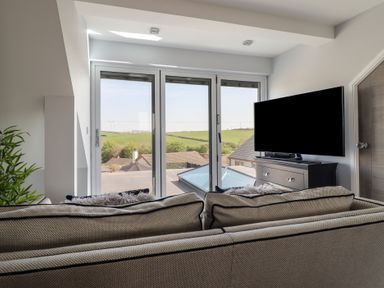 A seating area with a sofa facing a television and glass doors with a view of rooftops and green hills at Bronallt in Cemaes Bay