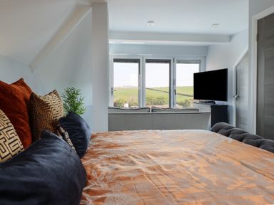 A bedroom with a bed and pillows facing a sofa and a television with windows showing a green field view at Bronallt in Cemaes Bay
