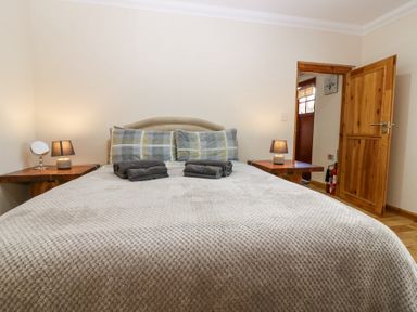 A bedroom with a bed covered with a textured blanket and folded towels two wooden bedside tables with lamps and a door open at Smiddy Bothy in Edzell