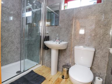 A bathroom with a glass shower enclosure a pedestal sink a toilet a trash can and a bath mat on a wooden floor at Smiddy Bothy Edzell