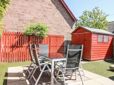 An outdoor patio with a glass table and six chairs near a red wooden shed and a red fence at Smiddy Bothy in Edzell