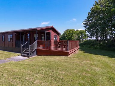 A wooden cabin with a deck and picnic table on a grassy lawn near trees at Lodge 9- Lamorna Tregolds near St Merryn
