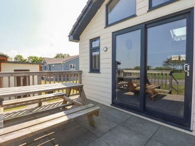 An outdoor patio with a wooden picnic table and benches next to a building with glass sliding doors and a window at Lodge 18 in St Merryn