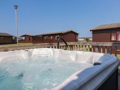 An outdoor hot tub on a deck with wooden cabins and a street lamp in the background at Lodge 18 in St Merryn