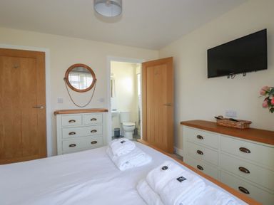 A bedroom with white bed linens and folded towels on the bed a dresser with drawers a wall-mounted TV and an open door leading to a bathroom at Lodge 18 in St Merryn