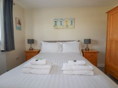 A bedroom with a bed with white linens and stacked towels on top two wooden bedside tables with lamps and a wooden wardrobe at Lodge 18 in St Merryn
