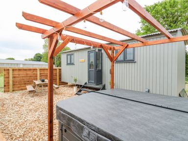An outdoor area with a wooden pergola over a hot tub a wooden fence a picnic table and a grey wooden cabin at Woodpecker in Lode near Bottisham