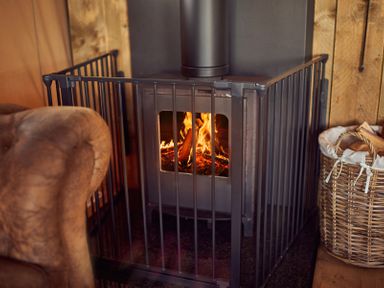 A wood burning stove with fire inside surrounded by a metal safety gate and a basket of firewood next to a brown armchair at Braeburn Lodge in Coxbridge near Glastonbury