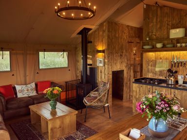 A living area with a brown sofa, wooden coffee table, armchair, wood stove, and kitchen with wooden cabinets and flowers in vases at Russet Lodge in Glastonbury