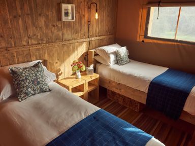 A bedroom with two single beds with white sheets and blue blankets separated by a wooden nightstand with a flower vase and tissues at Russet Lodge in Glastonbury