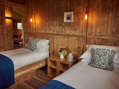 A bedroom with two single beds blue pillows wooden walls and a wooden nightstand with flowers and tissues at Russet Lodge in Glastonbury