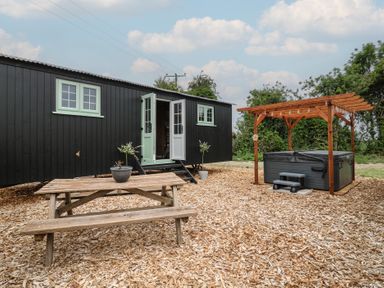 An outdoor area with a wooden picnic table a black caravan with mint green windows and doors and a hot tub with wooden pergola at The Heron in Lode near Bottisham