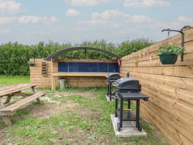 An outdoor area with two black barbecues a wooden picnic table a covered wooden bench and green plants at The Heron in Lode near Bottisham