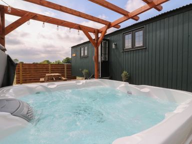 An outdoor hot tub with bubbling water under a wooden pergola next to a dark green cabin with two windows and a door at Barn Owl in Lode near Bottisham