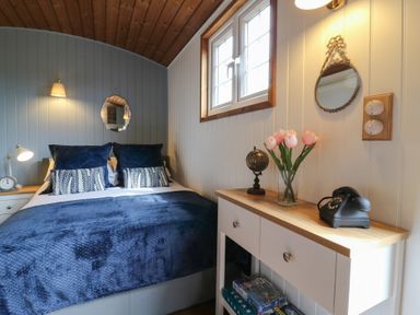 A bedroom with a bed blue bedding and pillows a wooden ceiling a side table with a clock a white cabinet with a globe flowers a vintage telephone and board games at Barn Owl in Lode near Bottisham