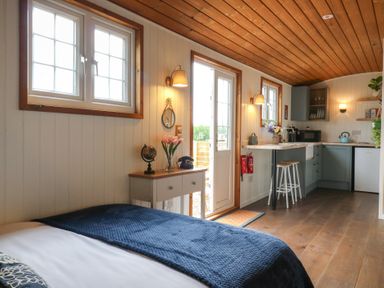A small room with a bed a side table with flowers and a vintage phone an open door leading outside and a kitchenette with a countertop and stools at Barn Owl in Lode near Bottisham