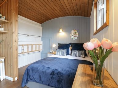 A bedroom with a bed with blue covers and pillows a wooden ceiling and a vase with pink flowers on a wooden table at Barn Owl in Lode near Bottisham