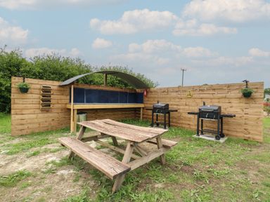 An outdoor picnic area with a wooden table benches two barbecues and a wooden shelter at Barn Owl in Lode near Bottisham