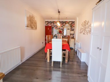 A dining room with a wooden dining table covered with a red tablecloth and wooden chairs in front of a floral wallpapered wall at 67 Pendre Avenue in Prestatyn