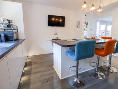 A kitchen with a counter and colorful bar stools a wall-mounted television and pendant lights at 67 Pendre Avenue in Prestatyn