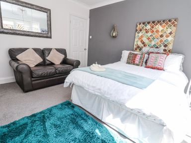 A bedroom with a bed with patterned cushions and a blue throw a brown leather sofa with two cushions a large mirror on the wall and a blue rug on the floor at 67 Pendre Avenue in Prestatyn